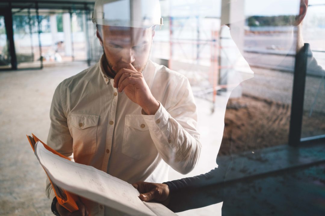 Two building engineers wearing safety hard hat discussing blueprint on construction site. Worker and inspector have meeting inside building under construction